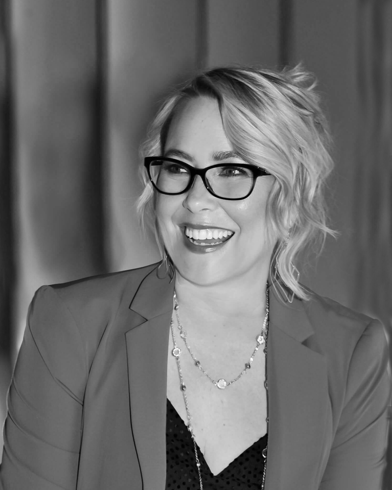 A woman with light, wavy, shoulder-length hair and dark-rimmed glasses.  She wears a medium blazer, dark v-neck top, and a shiny necklace.  The photo is black and white, with a softly blurred background.  She smiles toward the left of the camera and is illuminated from the right side.